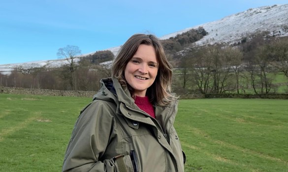 Headshot of woman wearing outdoor clothing stood in a green field with a snow-covered hill behind.